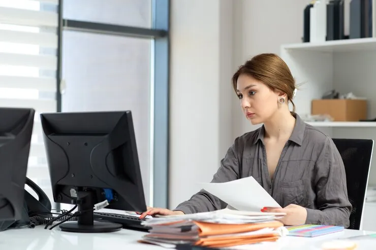 Accountant at the office with a stack of files, reading a document next to a computer